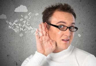 A man with glasses and a white sweater holds his hand to his ear, as if listening intently, with a cloud of floating letters and symbols around his head, against a grey background with hand-drawn clouds.