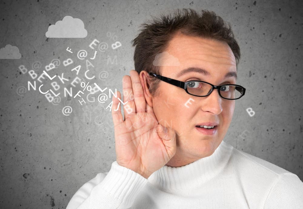 A man with glasses and a white sweater holds his hand to his ear, as if listening intently, with a cloud of floating letters and symbols around his head, against a grey background with hand-drawn clouds.