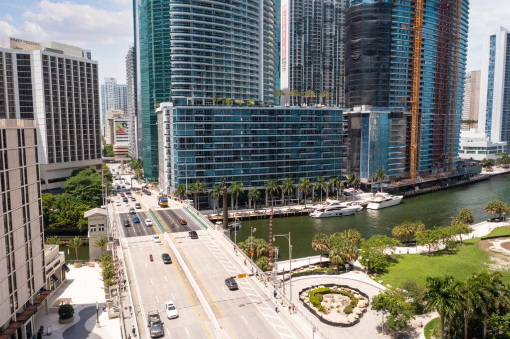 An aerial view of a busy bridge over the Miami River, surrounded by modern high-rise glass buildings, palm trees, and docked yachts in the Brickell neighborhood of Miami.