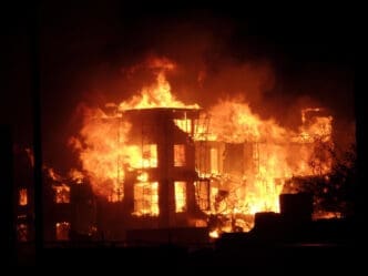 A large, multi-story building is completely engulfed in bright orange and red flames at night, with a large cloud of smoke rising into the air.