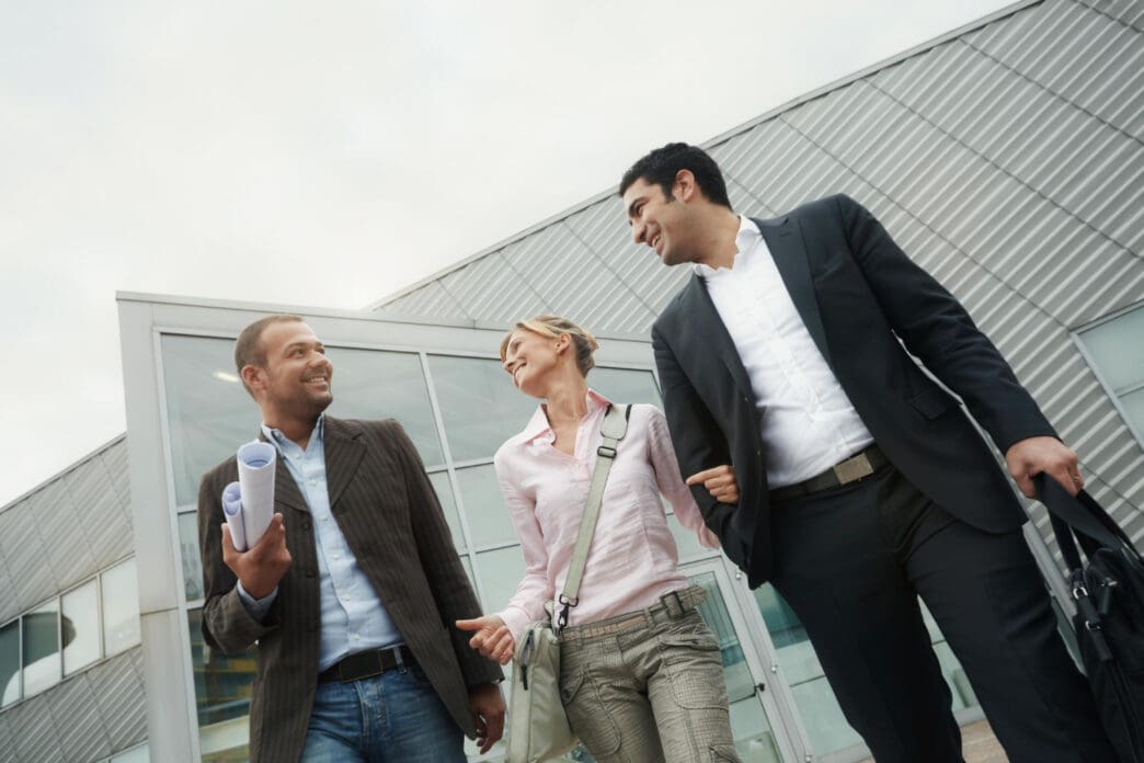 A low-angle photograph of three business colleagues, two men and one woman, walking and smiling as they talk outside a modern building with a corrugated metal facade.