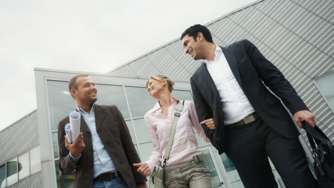 A low-angle photograph of three business colleagues, two men and one woman, walking and smiling as they talk outside a modern building with a corrugated metal facade.