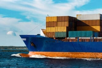 The front of a large, blue container ship with stacked containers on deck, sailing on the water