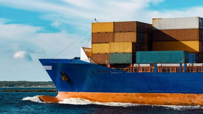 The front of a large, blue container ship with stacked containers on deck, sailing on the water