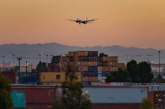A cargo plane flies low over a field of stacked containers at sunset.