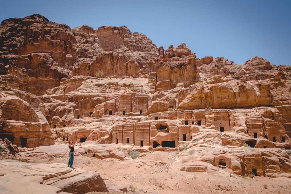 A wide view of ancient rock-carved tombs and buildings in Petra, Jordan, with a person standing in the foreground to show the scale.