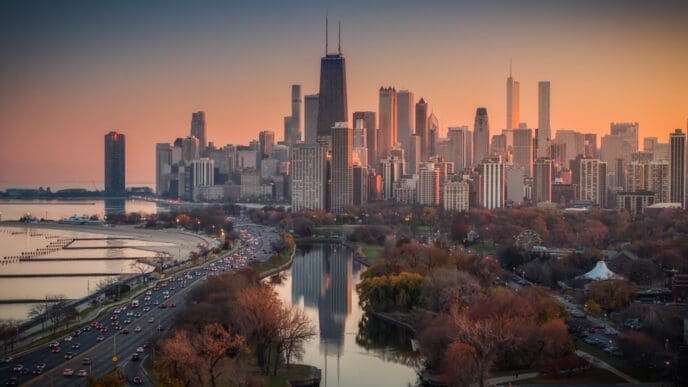 An aerial view of the Chicago city skyline at sunset, with a busy highway, a park, and a river reflecting the sky's colors in the foreground.