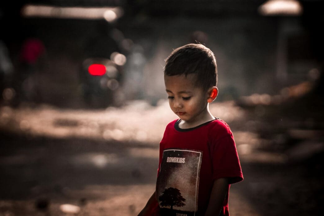A photograph of a young child in a red t-shirt looking down with a somber expression, with a blurred, dark and gritty background.