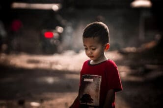 A photograph of a young child in a red t-shirt looking down with a somber expression, with a blurred, dark and gritty background.