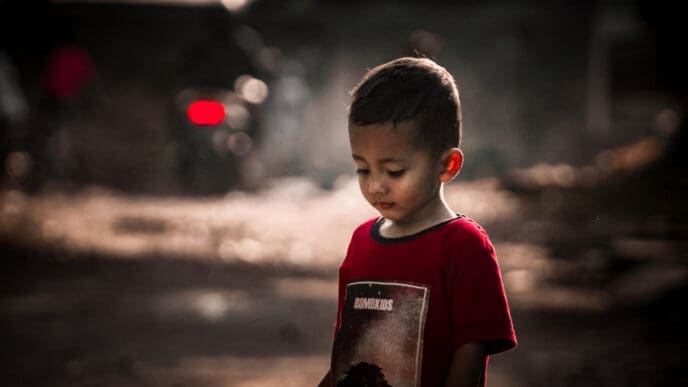 A photograph of a young child in a red t-shirt looking down with a somber expression, with a blurred, dark and gritty background.