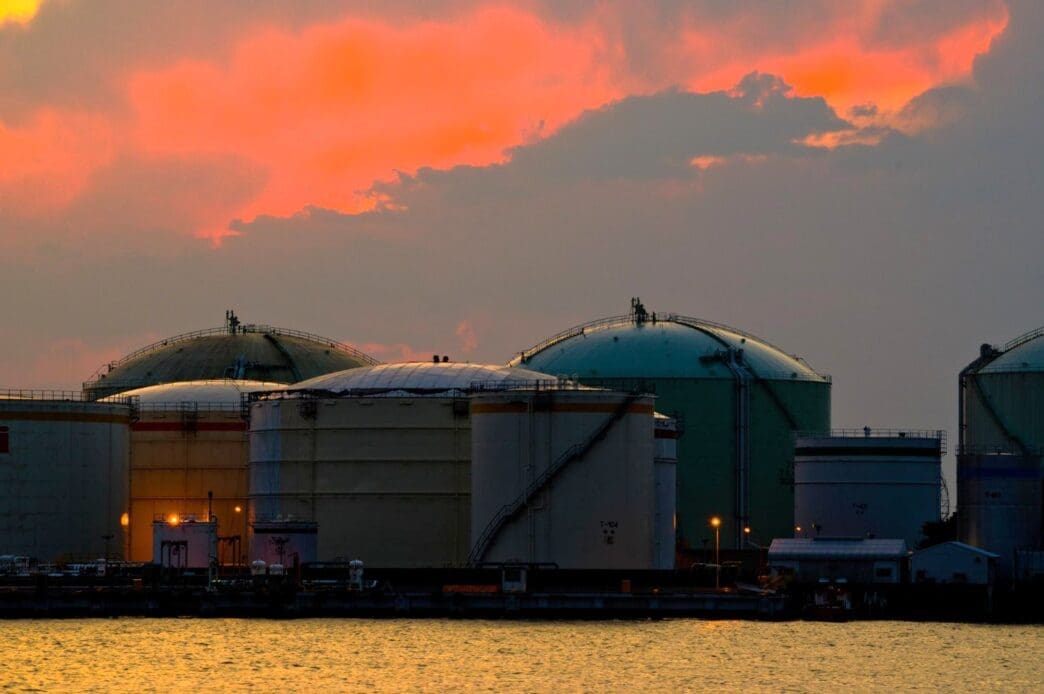 Large, domed industrial oil storage tanks are situated at a port, their silhouettes visible against a dramatic orange and pink sunset sky reflected on the water.
