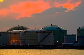 Large, domed industrial oil storage tanks are situated at a port, their silhouettes visible against a dramatic orange and pink sunset sky reflected on the water.