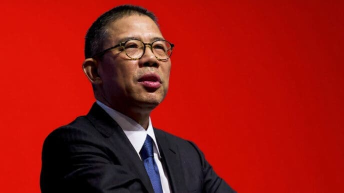 A close-up portrait of Chinese billionaire Zhong Shanshan, wearing a suit and glasses against a red background.