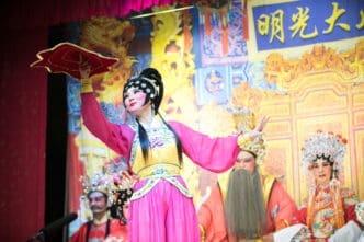 Performers in elaborate traditional costumes and theatrical makeup on a stage with a Chinese-themed backdrop.