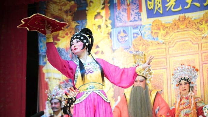 Performers in elaborate traditional costumes and theatrical makeup on a stage with a Chinese-themed backdrop.