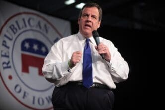 Former Governor Chris Christie speaks into a microphone, gesturing with a clenched fist, in front of a banner that says "REPUBLICAN OF IOWA."