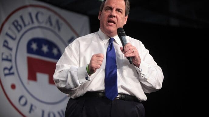 Former Governor Chris Christie speaks into a microphone, gesturing with a clenched fist, in front of a banner that says "REPUBLICAN OF IOWA."