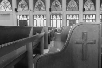 A black and white view of empty church pews with a cross carved into the last pew