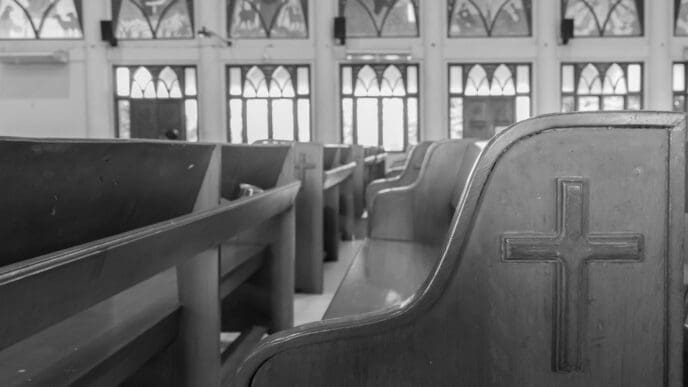 A black and white view of empty church pews with a cross carved into the last pew
