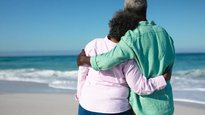 An older couple, seen from behind, embracing on a sandy beach while looking out at the ocean on a sunny day.