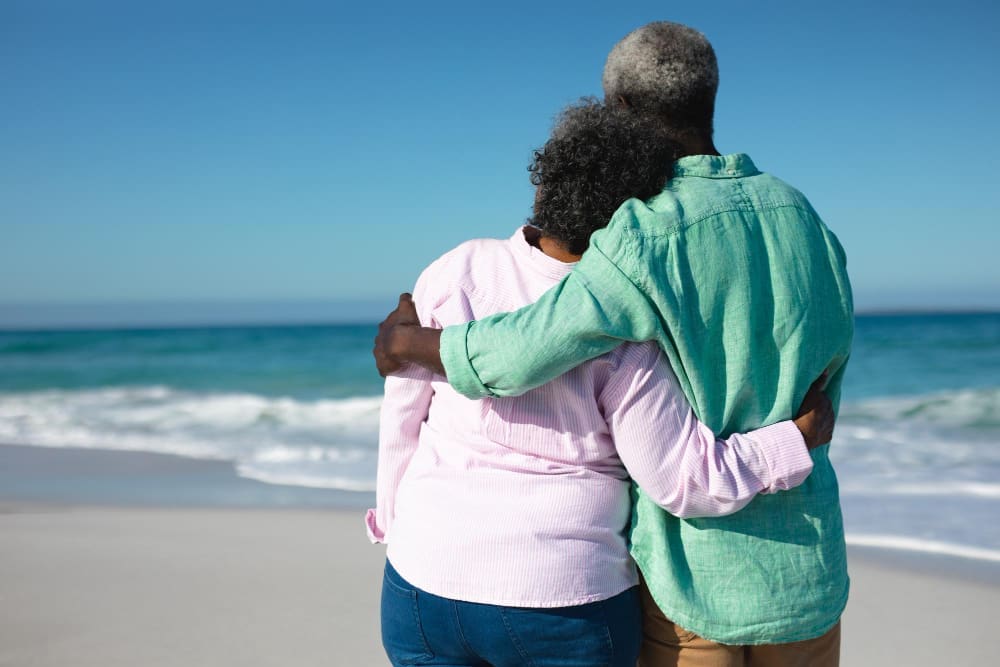 An older couple, seen from behind, embracing on a sandy beach while looking out at the ocean on a sunny day.