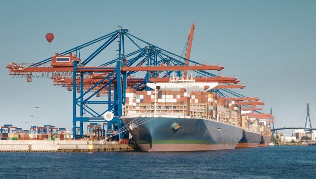 A large container ship is docked at a commercial harbor, with massive blue and orange gantry cranes overhead. A red hot air balloon is visible in the distance under a clear sky.