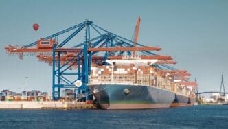 A large container ship is docked at a commercial harbor, with massive blue and orange gantry cranes overhead. A red hot air balloon is visible in the distance under a clear sky.