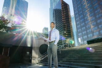 A businessman with papers smiles while standing in front of a city skyline with sun flare