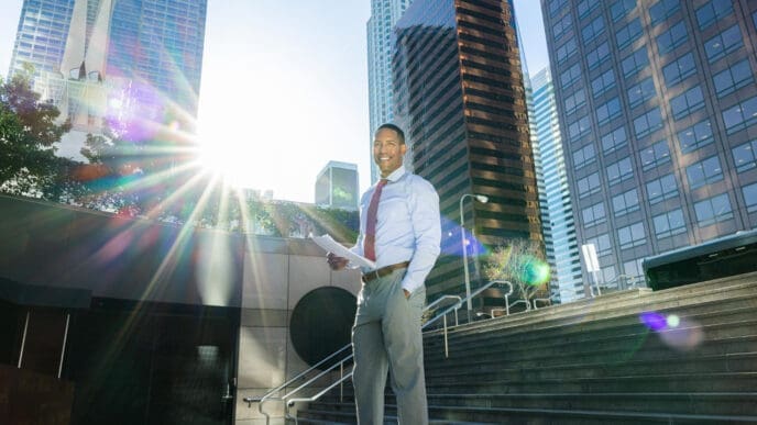 A businessman with papers smiles while standing in front of a city skyline with sun flare