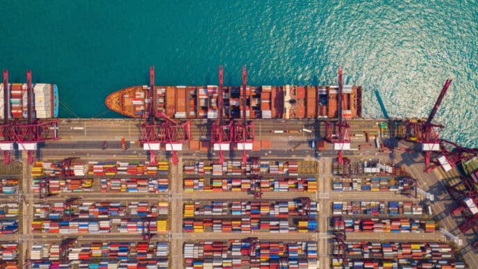 An aerial view of a busy shipping port, showing a container ship docked at the pier and thousands of multi-colored shipping containers stacked on the ground.