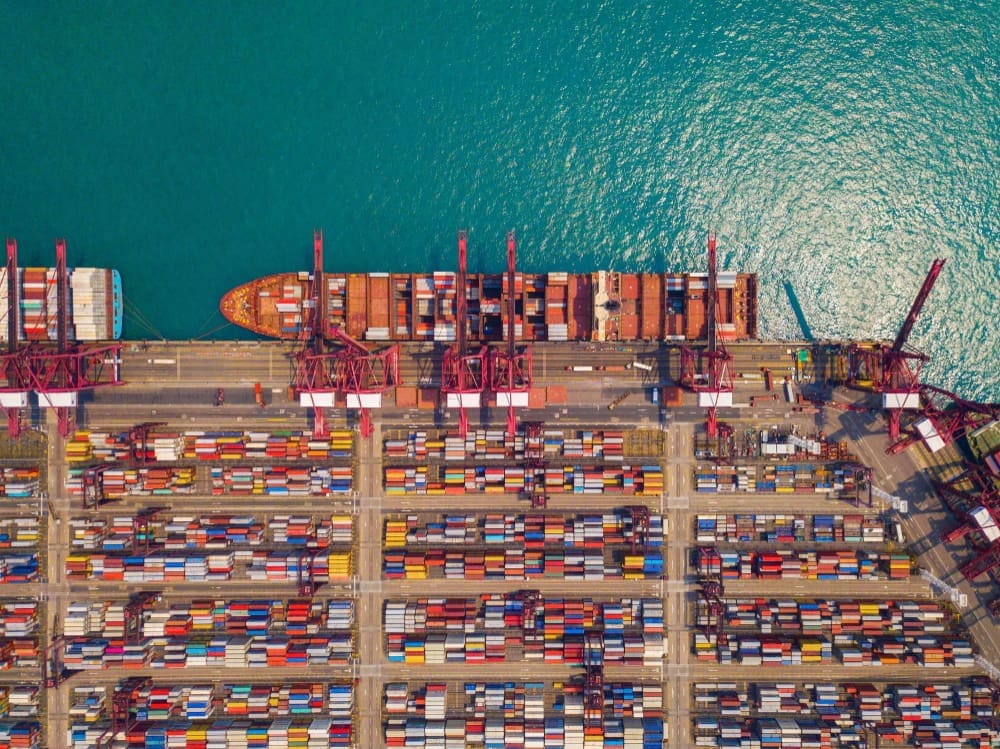 An aerial view of a busy shipping port, showing a container ship docked at the pier and thousands of multi-colored shipping containers stacked on the ground.