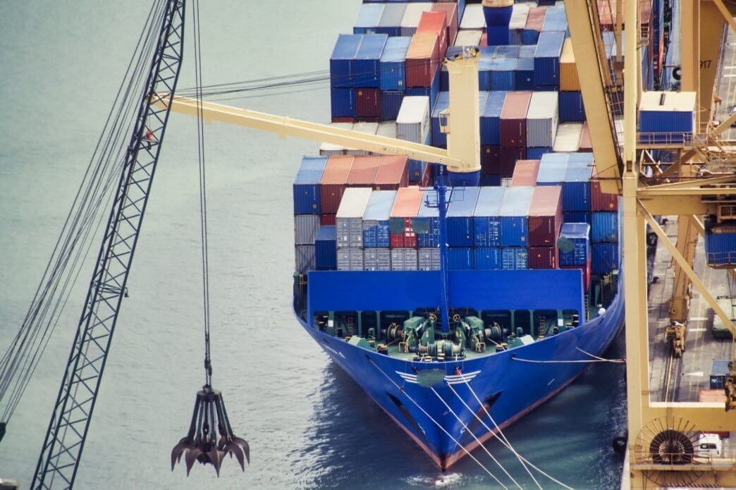 A high-angle view of a large blue container ship loaded with colorful containers, docked at a port and positioned next to a large crane.