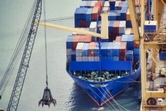 A high-angle view of a large blue container ship loaded with colorful containers, docked at a port and positioned next to a large crane.