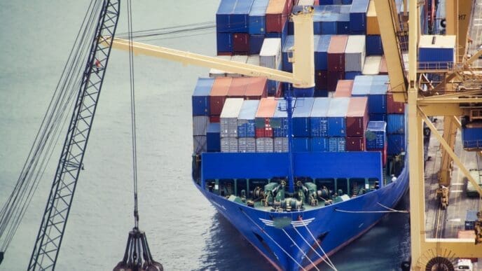 A high-angle view of a large blue container ship loaded with colorful containers, docked at a port and positioned next to a large crane.