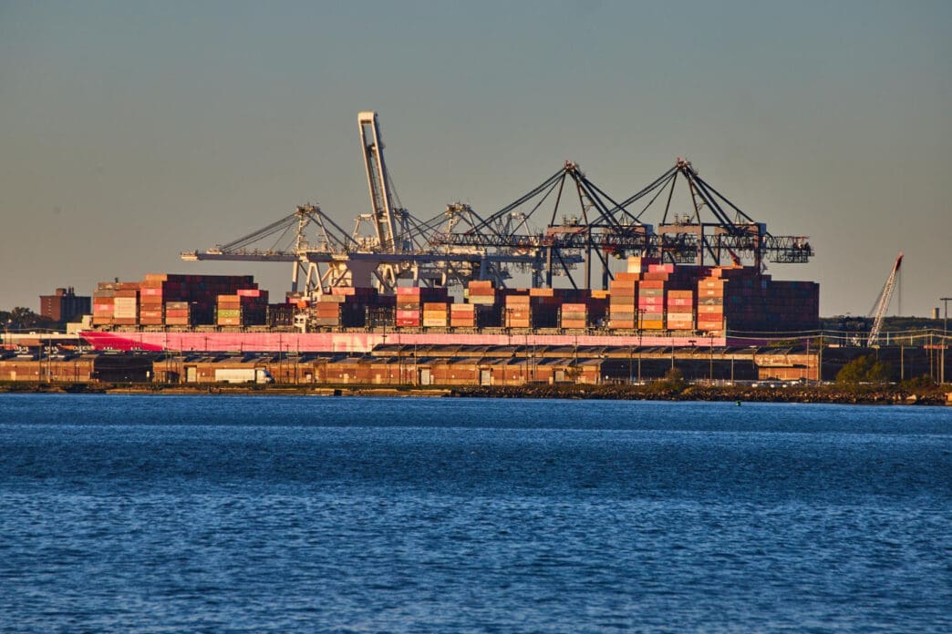 A large container ship loaded with shipping containers is docked at a port in New York City, seen in the golden hour light.