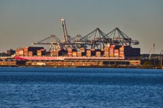 A large container ship loaded with shipping containers is docked at a port in New York City, seen in the golden hour light.
