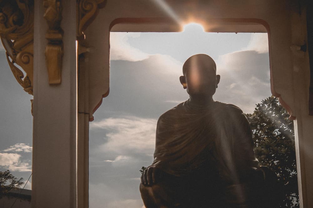 A silhouetted statue of a meditating figure, likely a Buddhist monk, framed by an archway with a sunburst and clouds behind it.
