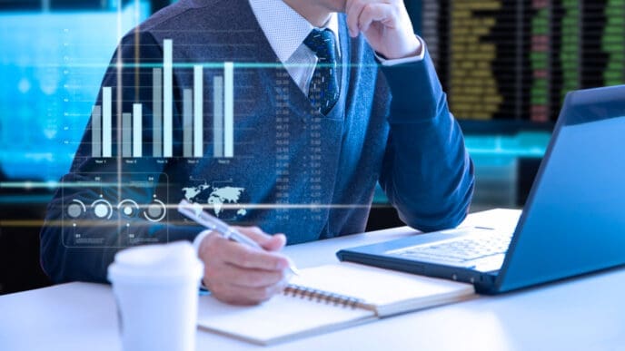 A person in a sweater vest and tie, working at a desk with a laptop and notebook, with a holographic overlay of digital charts and graphs, representing the automation of corporate finance tasks.