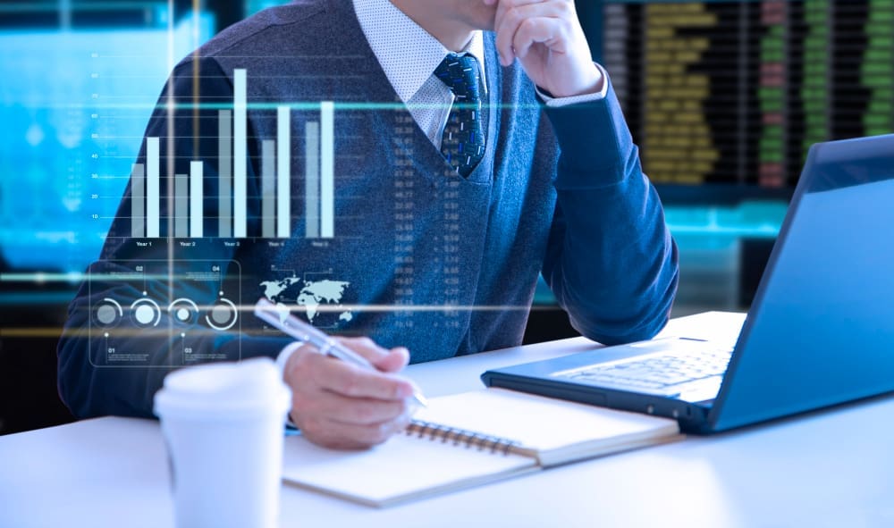 A person in a sweater vest and tie, working at a desk with a laptop and notebook, with a holographic overlay of digital charts and graphs, representing the automation of corporate finance tasks.