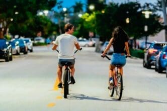 A young couple, seen from behind, is biking down a street in Miami Beach at dusk, with the man on the left and the woman on the right, surrounded by parked cars and streetlights.
