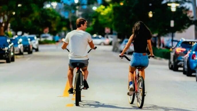 A young couple, seen from behind, is biking down a street in Miami Beach at dusk, with the man on the left and the woman on the right, surrounded by parked cars and streetlights.