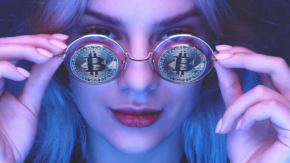 A woman holding up round glasses with Bitcoin coins for lenses, looking at the camera under a blue and purple light.