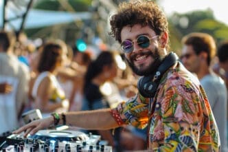 A smiling DJ with a beard and sunglasses, wearing a brightly colored shirt and headphones, with his hand on a mixing board at a daytime party.