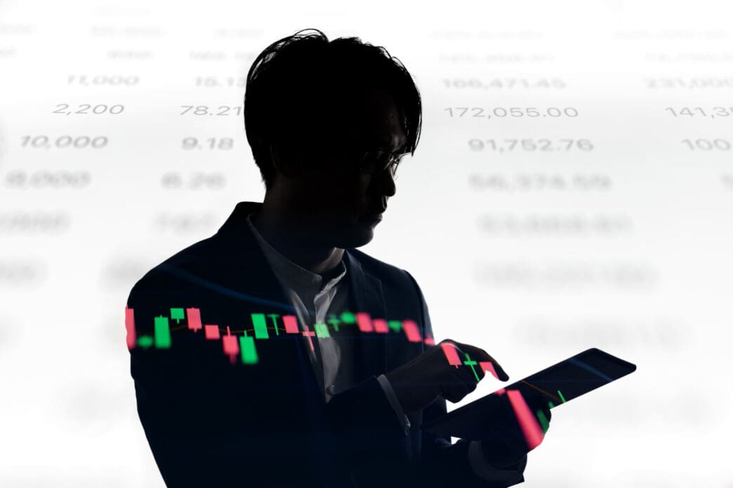 The silhouette of a person in a suit is shown looking at a tablet, with a glowing financial candlestick chart overlaid on both the tablet and their body.