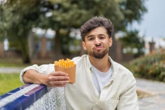 A young indian man with a sad expression holds a container of french fries while sitting outdoors.