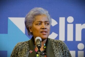 Donna Brazile, a woman with short gray hair, speaks into a microphone at a campaign event with a blue background and a blurred logo.