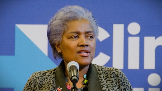 Donna Brazile, a woman with short gray hair, speaks into a microphone at a campaign event with a blue background and a blurred logo.