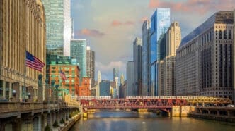The Chicago River runs through the city, with a red metal bridge and an elevated train crossing it, surrounded by a dense skyline of skyscrapers.