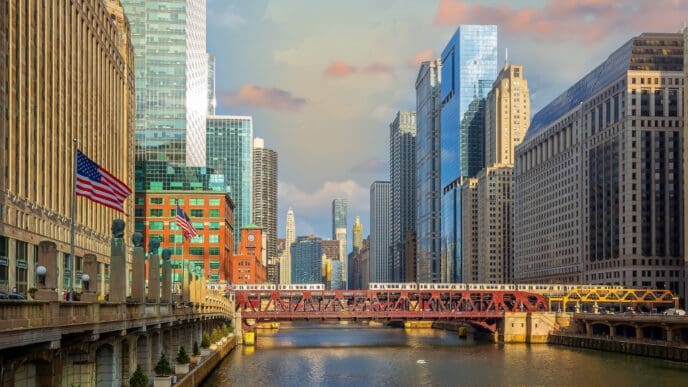 The Chicago River runs through the city, with a red metal bridge and an elevated train crossing it, surrounded by a dense skyline of skyscrapers.