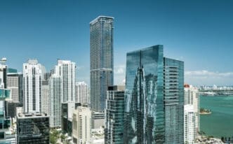 An aerial view of the dense, modern Miami skyline, with numerous tall skyscrapers of glass and white facades, overlooking the blue-green bay with distant land and a port, all under a clear blue sky.